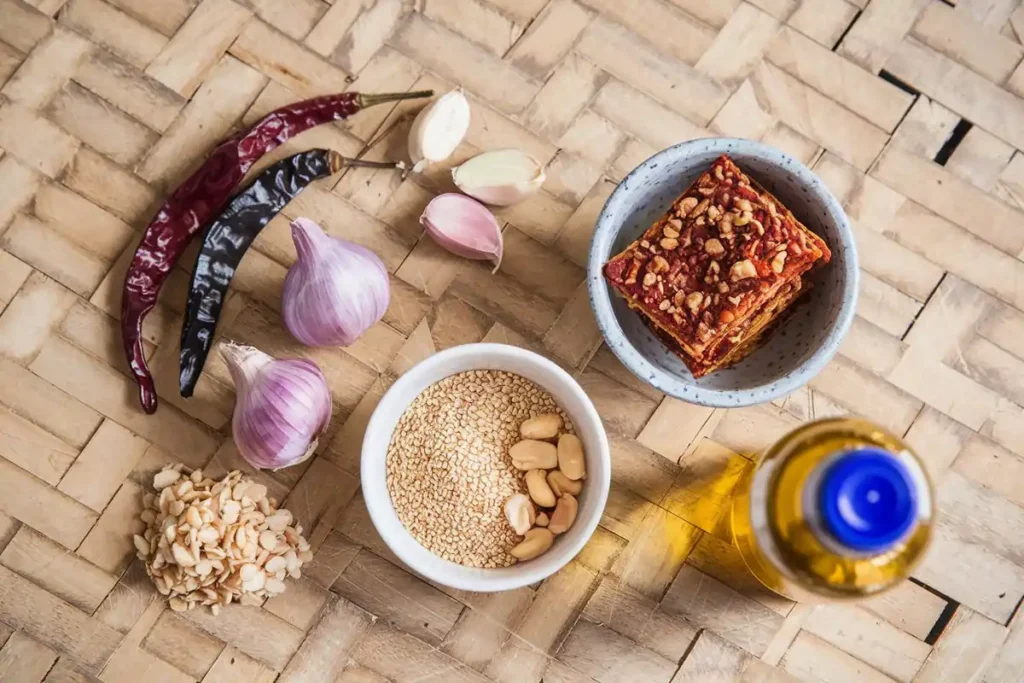 Ingredients for Kari Kari Chili Crisp, including dried chilies, garlic, shallots, sesame seeds, peanuts, and cooking oil, arranged on a wooden board.