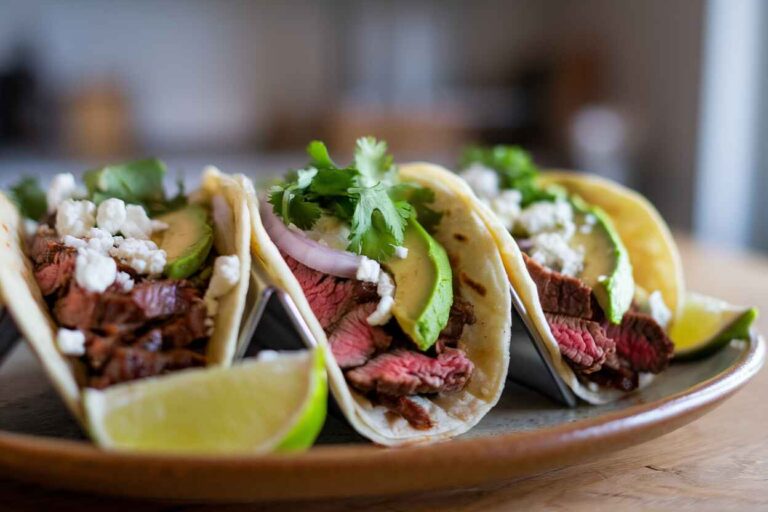 Three chipotle steak tacos on warm corn tortillas, topped with chopped onions, cilantro, avocado, and crumbled queso fresco.