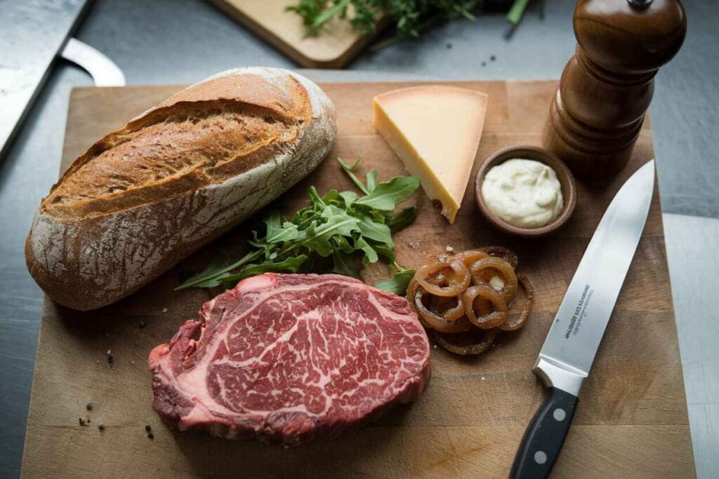 A flat-lay of premium steak, freshly baked bread, cheese, sauces, and toppings for making a steak sandwich recipe.