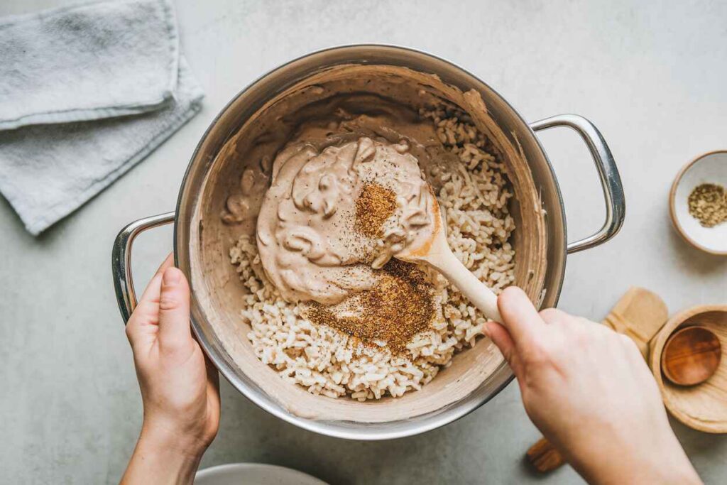 A bowl filled with uncooked long-grain rice, cream of mushroom soup, chicken broth, and seasonings being mixed.